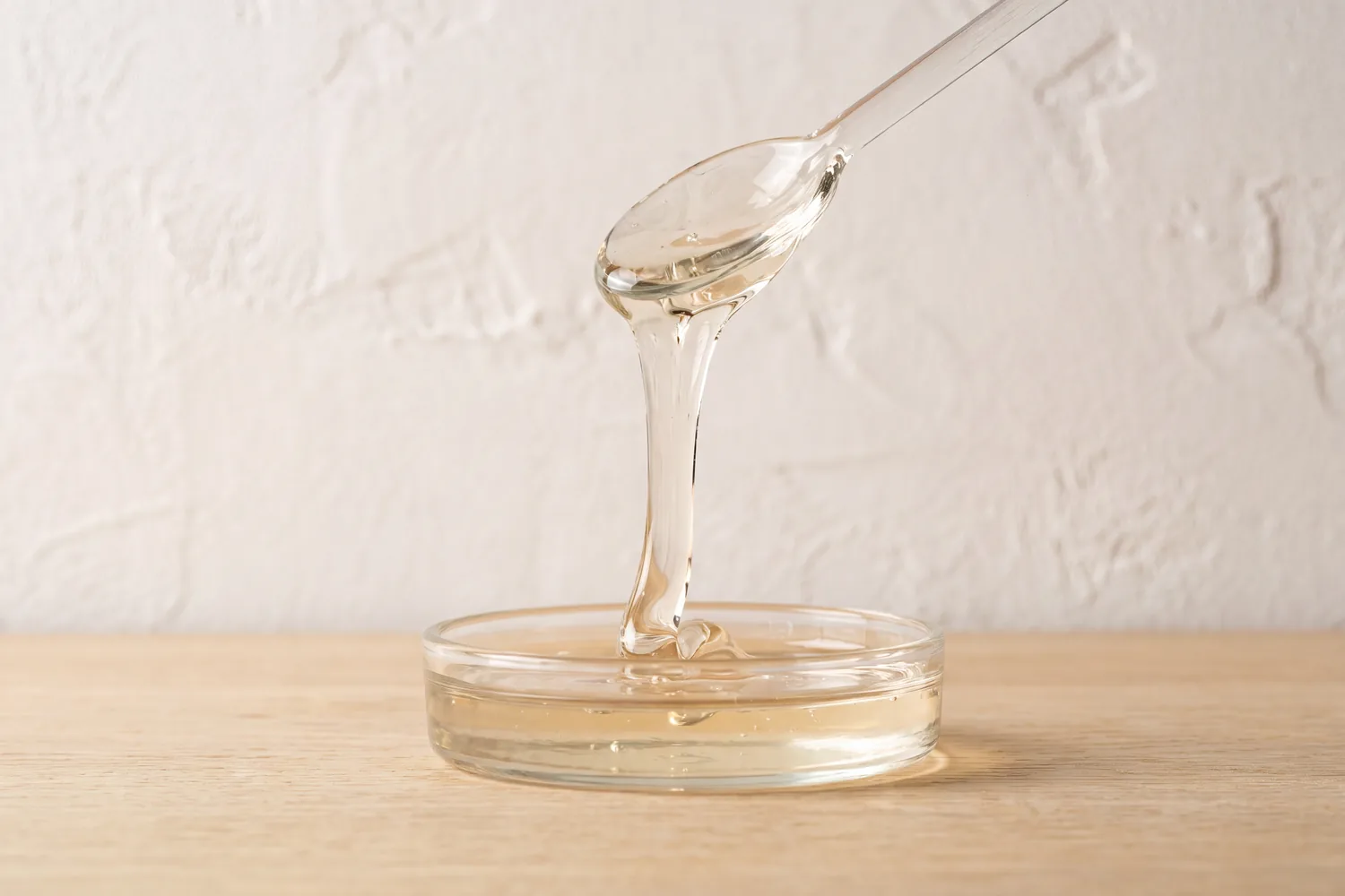 A glass spoon letting a slow ribbon of clear glycerin fall into a shallow glass dish.